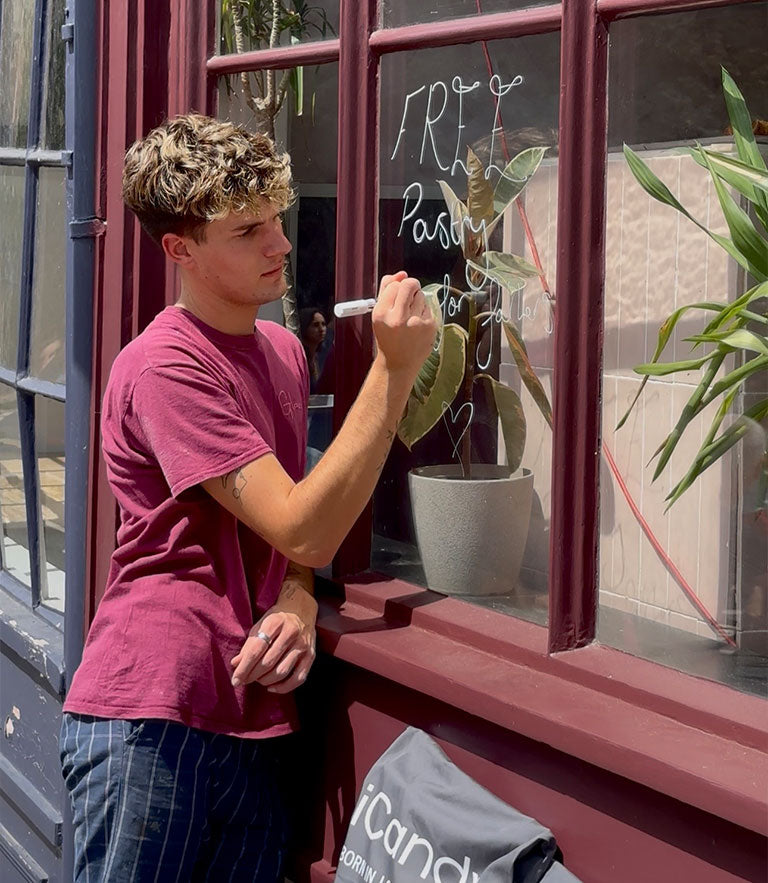 A young man with curly blonde hair wearing a maroon t-shirt and striped pants stands by a window, writing on the glass with a white marker. A gray tote bag with the brand name 'iCandy' is resting on the window sill below him.